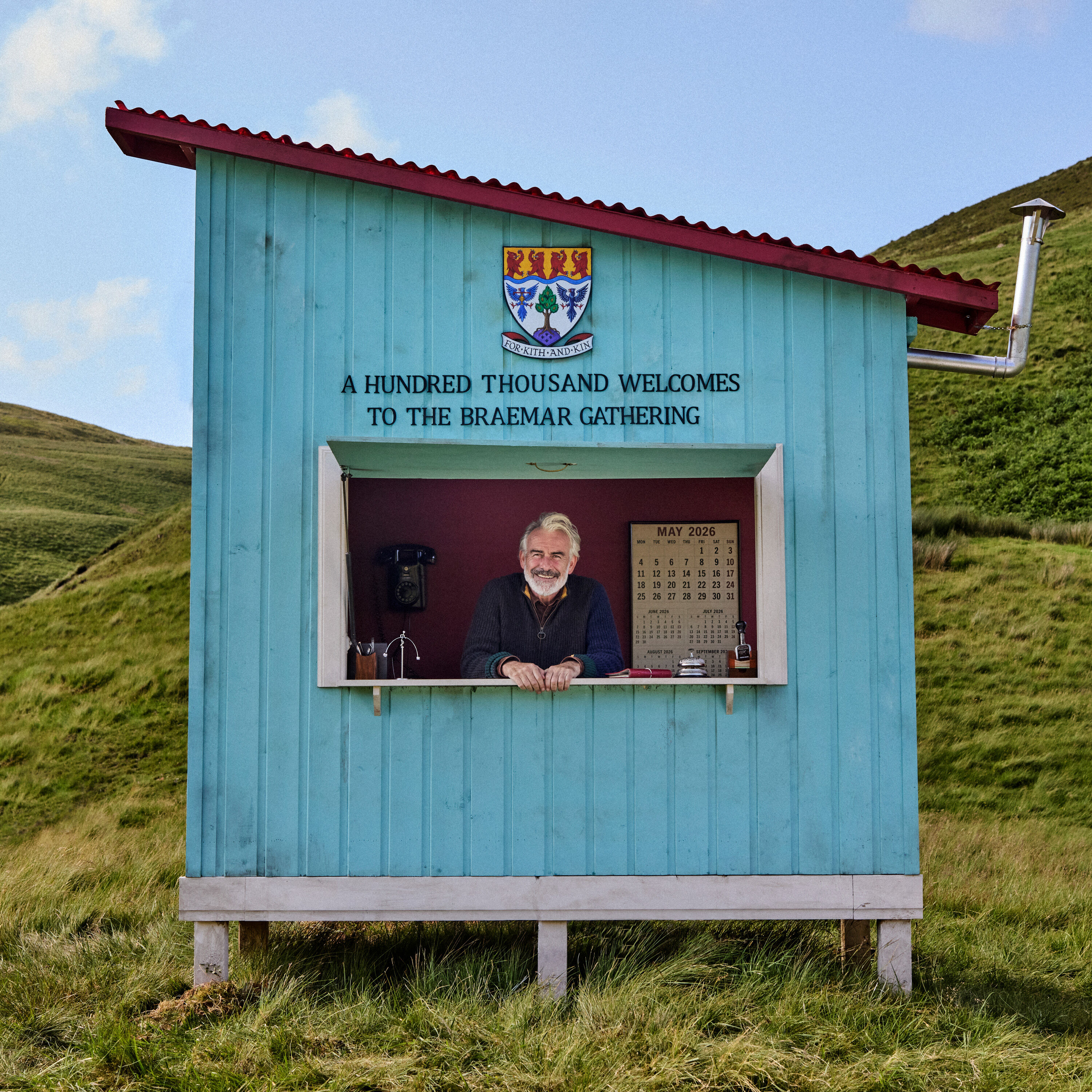 Man standing in blue hut with sign that reads 'A Hundred thousand welcomes to the Braemar gathering'