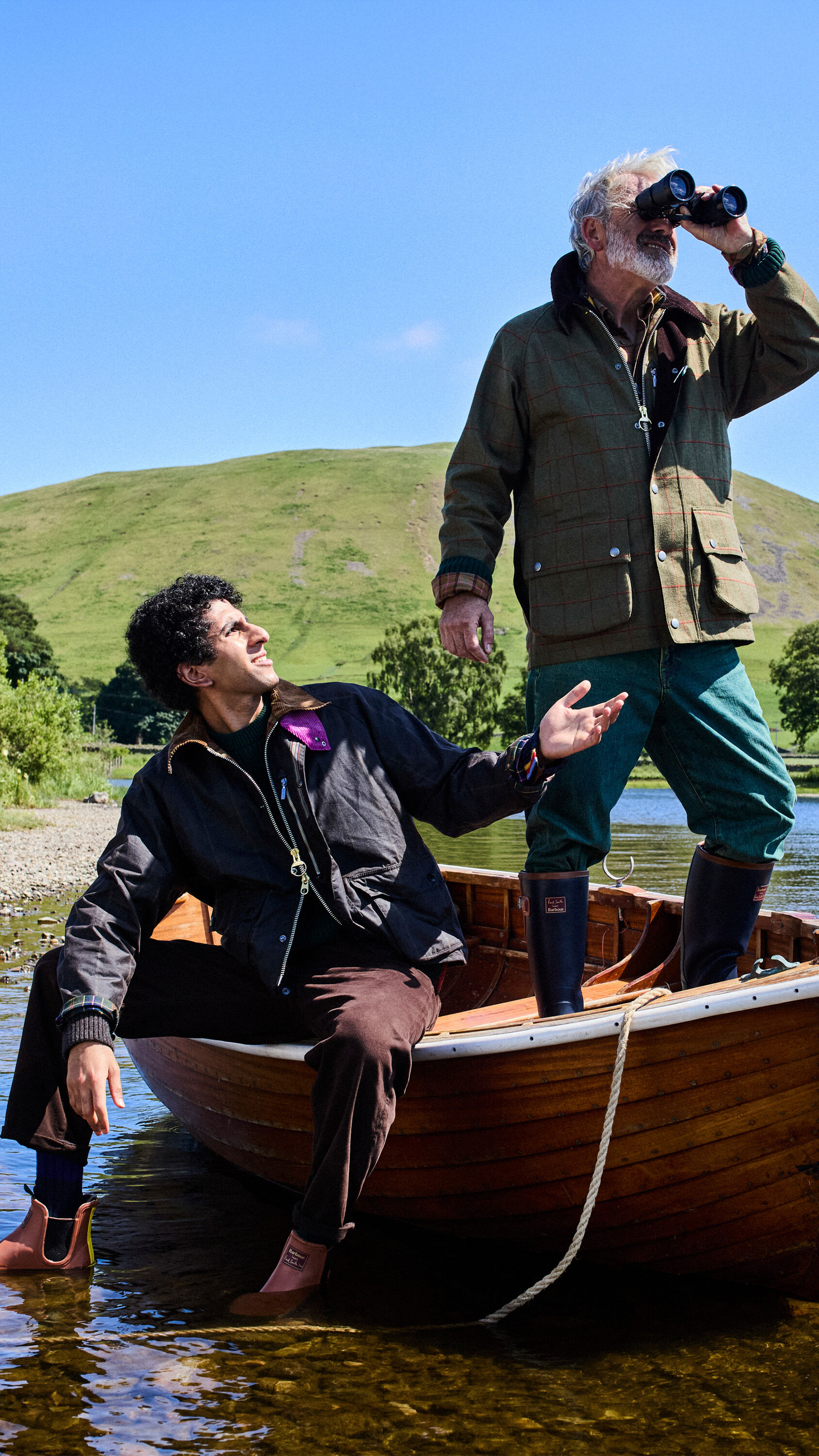 Two men standing in a baot, one wearing the Barbour x Paul Smith Transport Windowpane Wax Jacket in rustic/bark, the other man wearing the Barbour x Paul Smith Beaufort Wool Jacket in olive window pane